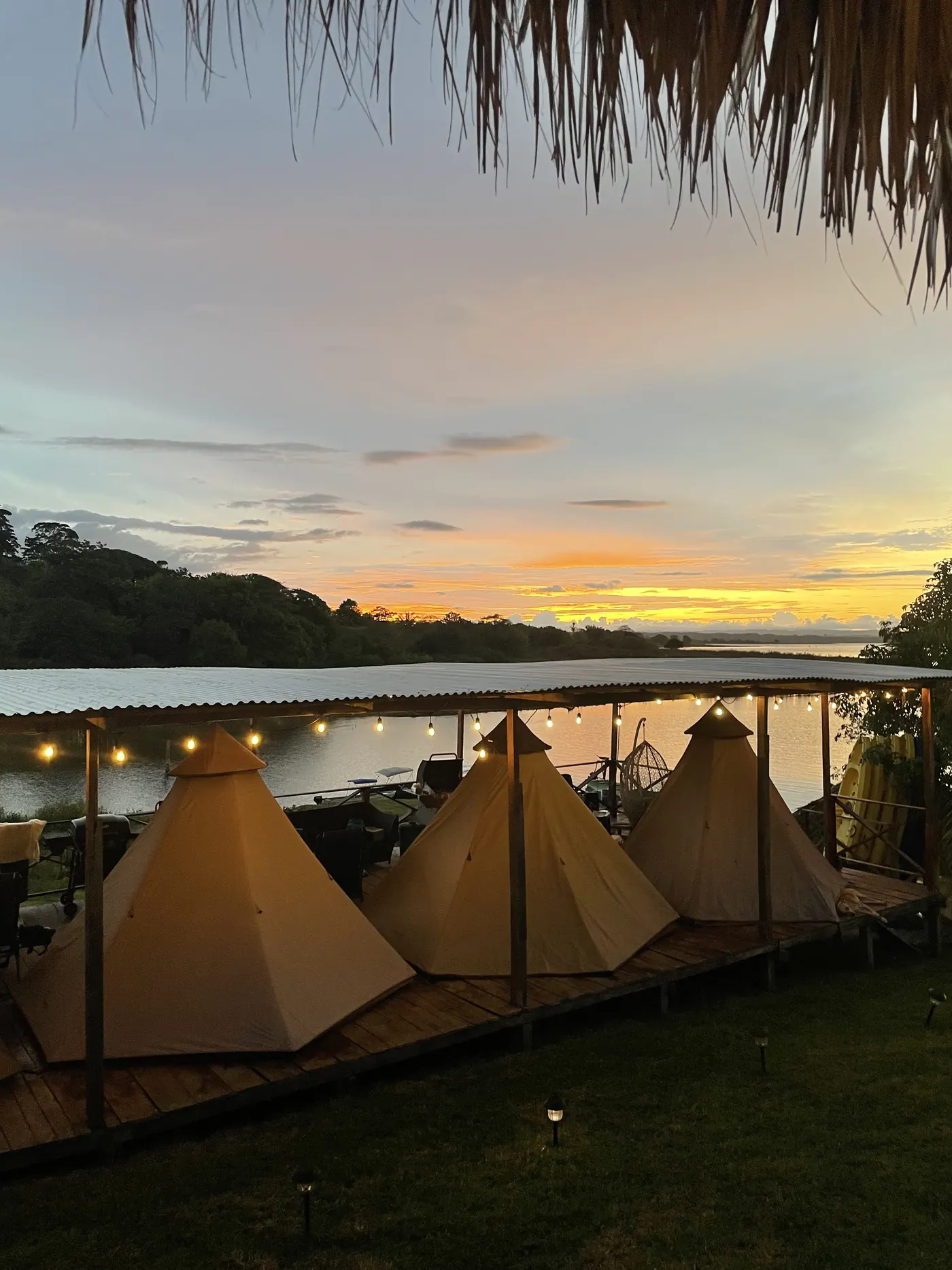Tres carpas cónicas sobre un deck de madera junto al lago, iluminadas con luces cálidas al atardecer