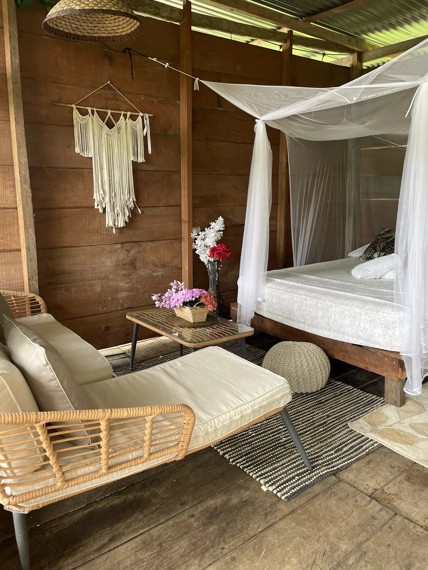 Cama con dosel de mosquitero junto a un sofá y otomana de ratán, con un colgante de macramé en la pared de madera