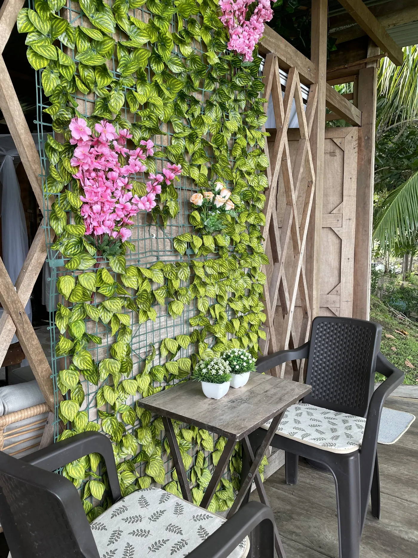 Rincón con muro de enredaderas y flores rosadas, y una mesa pequeña con dos sillas para desayunar afuera.