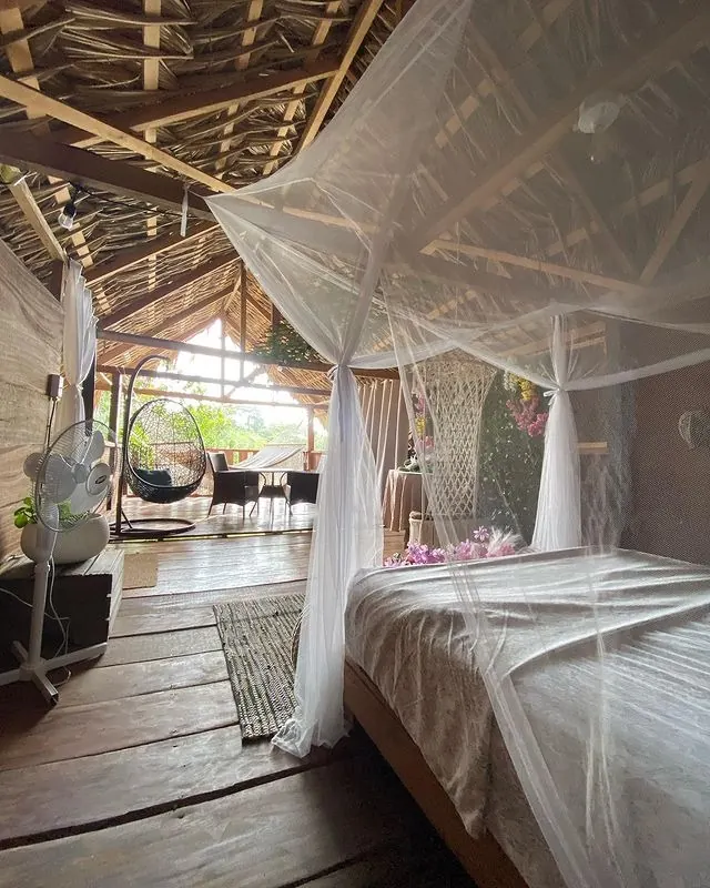 Cama con mosquitero cubierta por un techo bajo de palma con vigas de madera a la vista, y una silla colgante negra tipo huevo junto al comedor al fondo