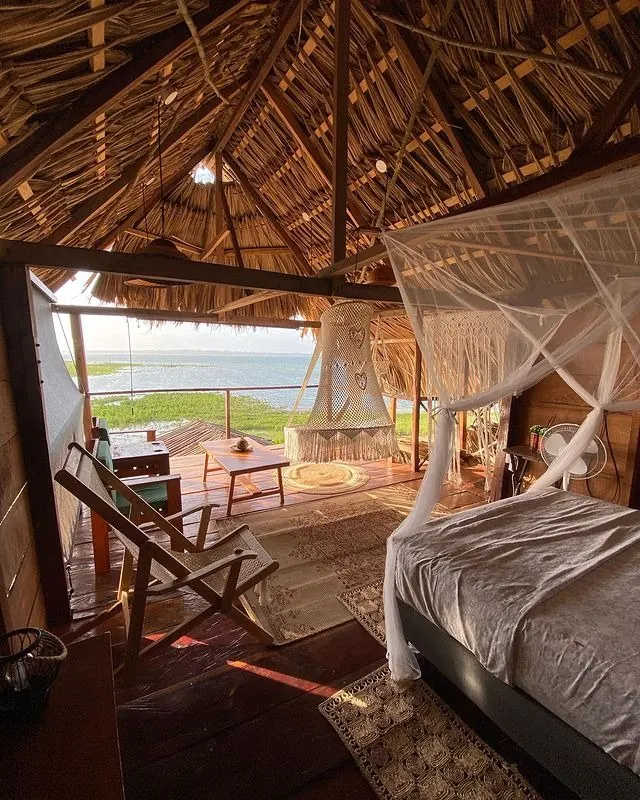Cama con mosquitero bajo un techo de palma en punta, con una lámpara colgante de macramé junto al deck que se abre directo al lago, en luz dorada de atardecer