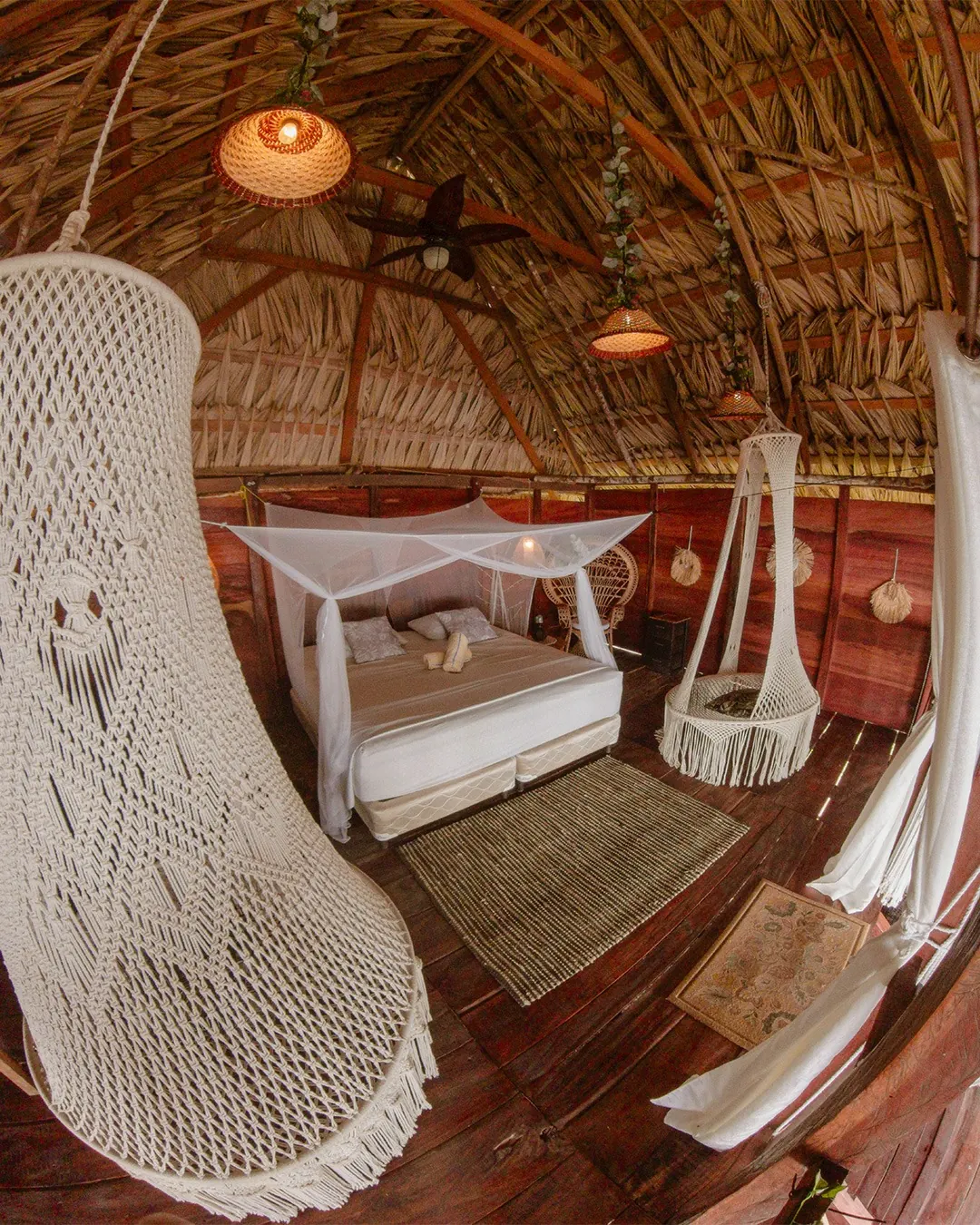 Interior de un solo nivel con techo de palma abovedado, cama con mosquitero a ras de piso y una gran silla colgante de macramé en primer plano