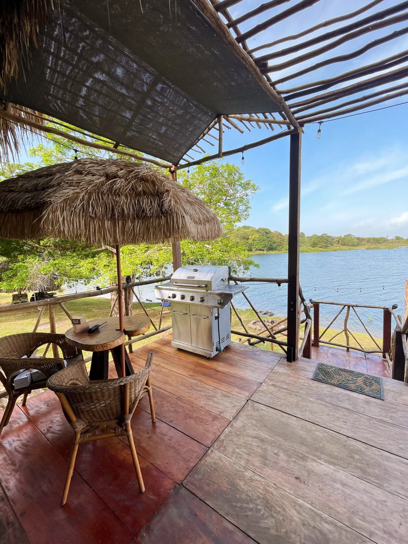 Comedor exterior con parrilla de gas, bajo una sombrilla de palma y vista al lago.