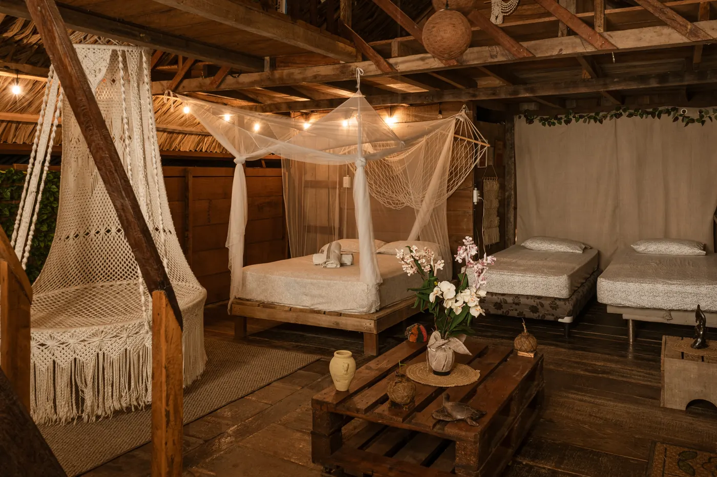 Cama principal con dosel de mosquitero, dos camas individuales a la derecha, silla colgante de macramé a la izquierda y la estructura de madera del altillo visible arriba