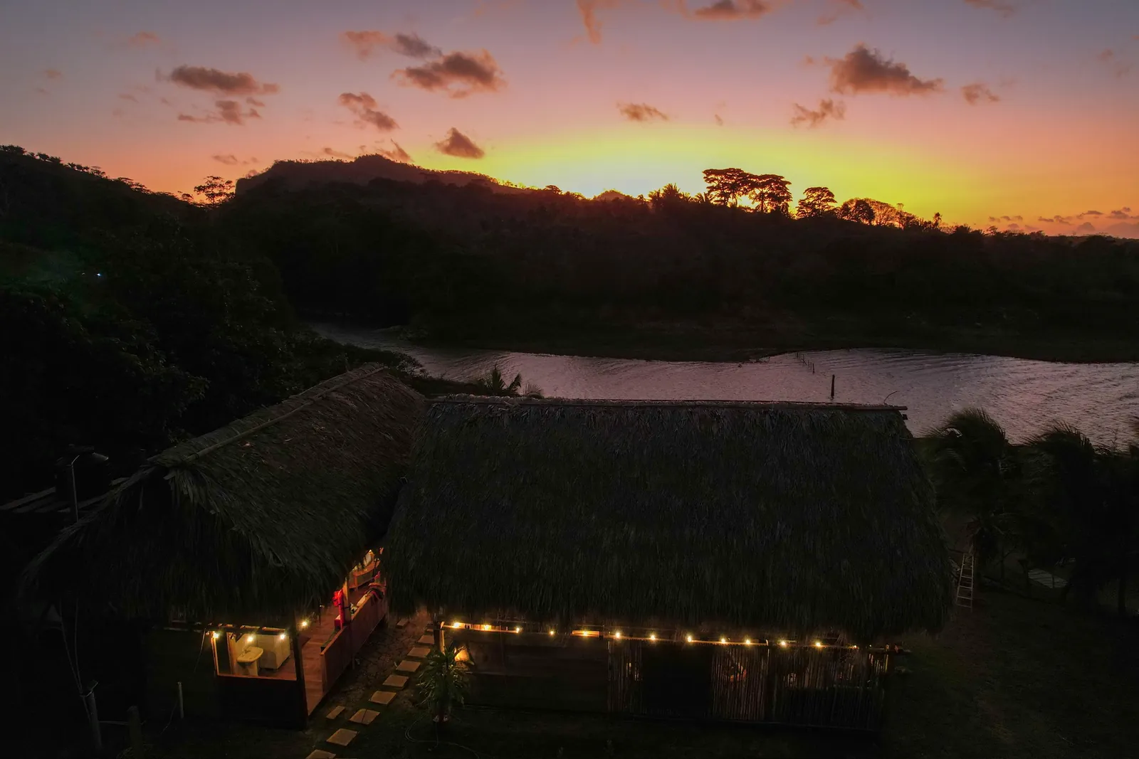 El ecolodge de noche desde el aire: techos de penca, luces encendidas y el cielo en tonos naranjas