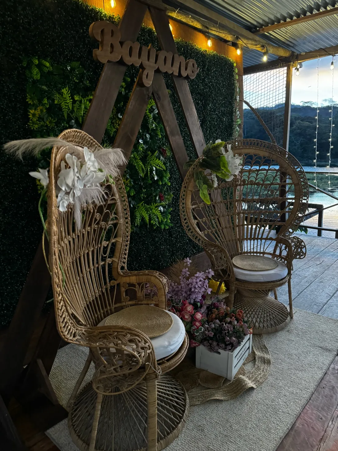 Montaje de celebración al atardecer: dos sillas de mimbre con flores, el letrero de Bayano y luces colgantes, con el lago al fondo